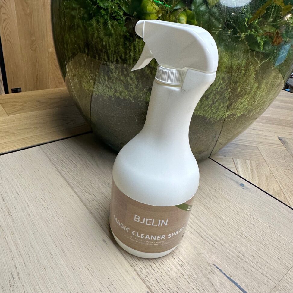 Spray bottle labeled 'Bjelin Magic Cleaner Spray' on a wooden floor with a decorative plant in the background.