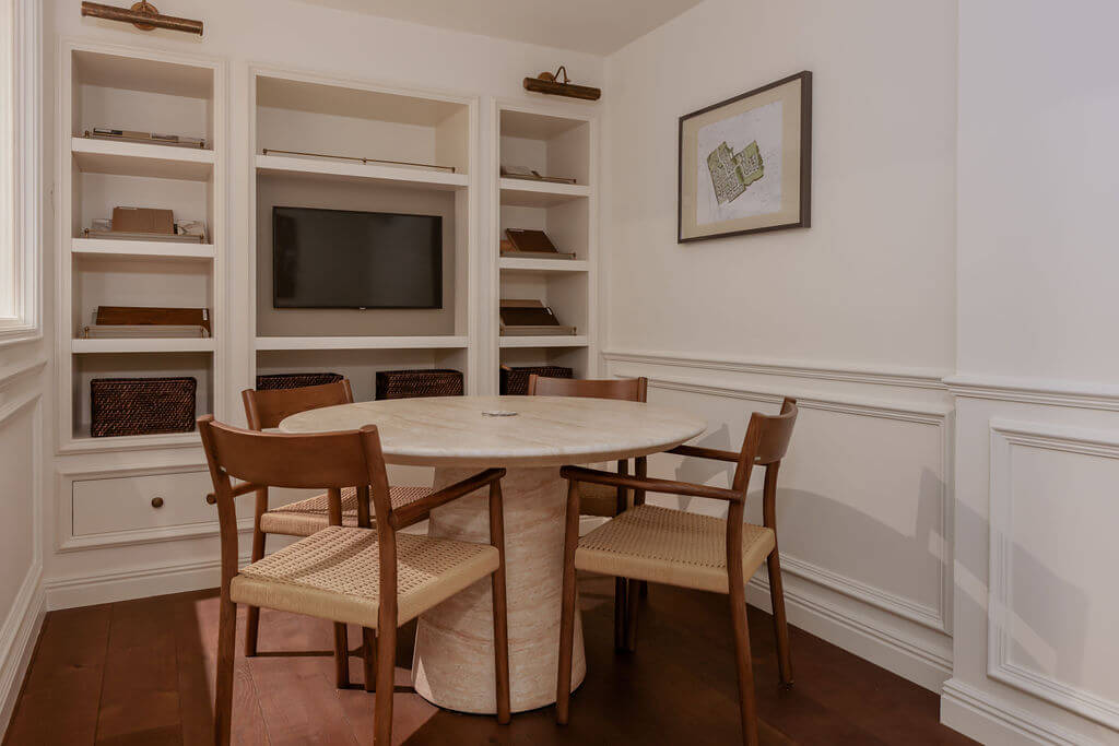 A round meeting table with a light stone top and a textured pedestal base, surrounded by four wooden chairs with woven seats in a space with v4 wood flooring