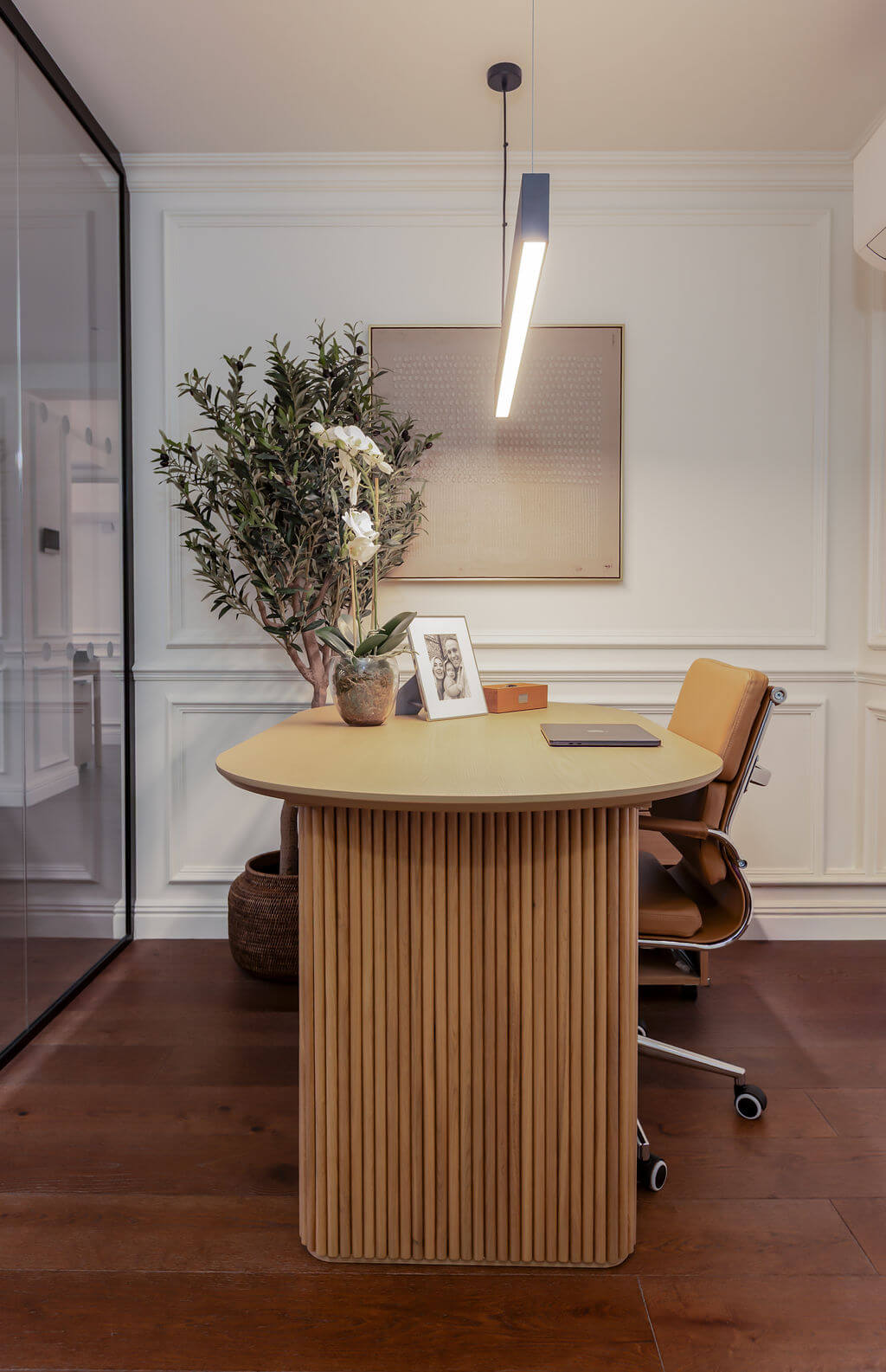 A modern wooden desk with a fluted slatted pedestal base and a rounded top, styled in a bright office with dark wood floors