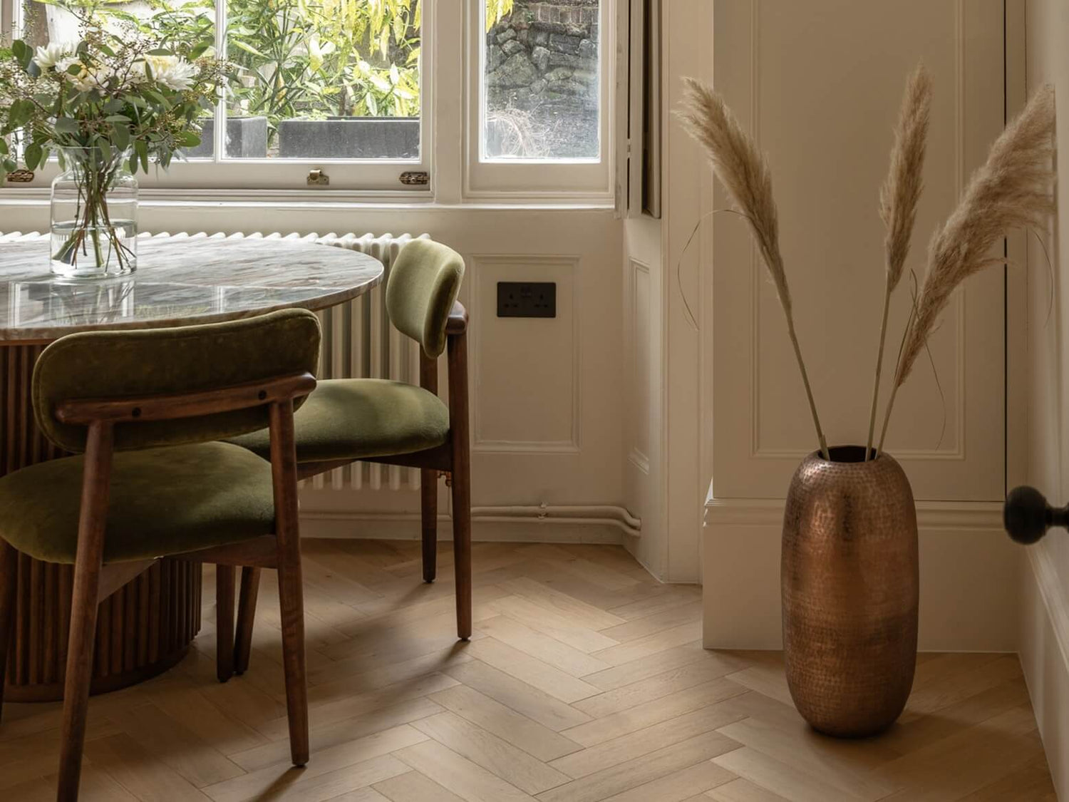 Dining area with wooden table, chairs, and a vase with dried plants on a herringbone floor.