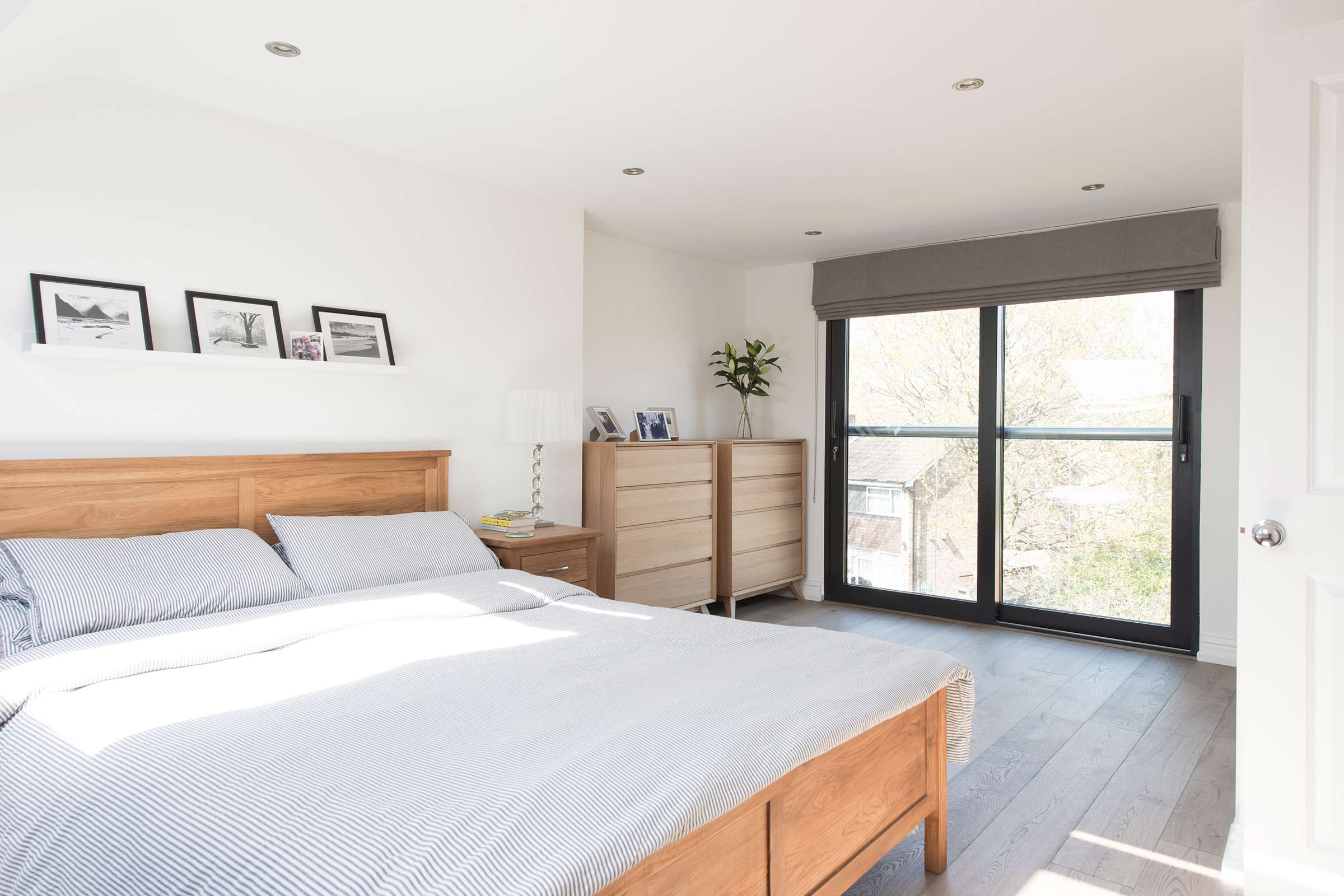 A light, airy bedroom with floor to ceiling windows, white walls, wooden furniture, and frozen umber wood flooring