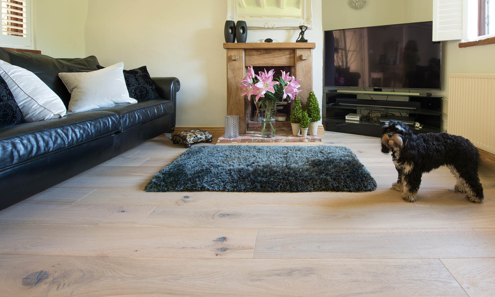 a cottage living room with a small dog and oak wood floors, leather furniture and pink lilies on the fireplace mantle